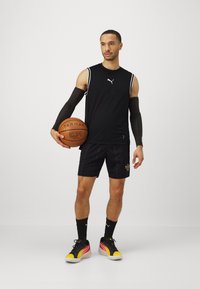 Top atletico nero senza maniche con strisce bianche, pantaloni corti neri con motivo testurizzato, maniche nere per il braccio e scarpe da basket multicolori.