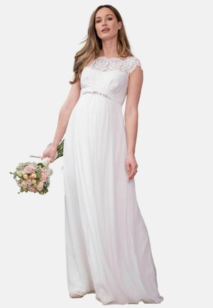 Kvinde i hvid blonde bryllupskjole, der holder en buket med pink og hvide blomster, stående mod en ensfarvet lys baggrund.