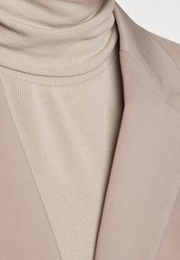 Detailní záběr béžového žebrovaného roláku naskládaného pod límcem světle hnědého blejzru.