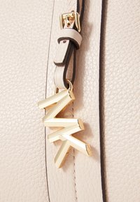 Bandoulière de sac à main beige texturée avec une petite boucle beige et un charm en métal doré en forme des lettres Z, M et K suspendu.