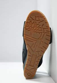 Semelle d'une bottine Timberland noire montrant une bande de roulement détaillée en caoutchouc orange avec le logo de l'arbre et des motifs géométriques.