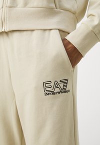 Bež trenirka iz bombaža z rebrastimi manšeti, ki ima sprednji zadrgo in črn logotip "EA7 EMPORIO ARMANI" na žepu. Mehek občutek.
