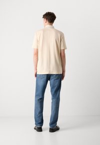 Beige kortärmad polo med krage, tillsammans med ljusblå raka jeans och svarta skor. Slät tygtextur.