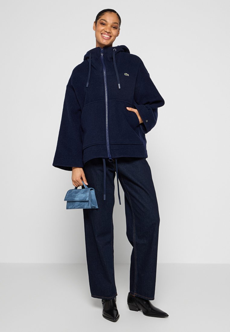 Marineblaue Kapuzenfleecejacke mit einem Frontreißverschluss, zwei Taschen und weiten Ärmeln. Kombiniert mit dunkelblauen Jeanshosen und einer kleinen blauen Handtasche.