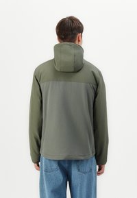 Grøn hættetrøje med et tofarvet design. Glat tekstur, justerbar hætte og let glans. Samlet med blå jeans og en ensfarvet baggrund.