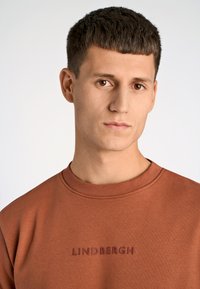 Felpa di cotone marrone con scollo rotondo, caratterizzata da un logo 'LINDHERGH' testurizzato di un marrone più scuro sul davanti. Tessuto liscio e forma aderente.