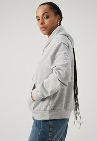 Gray hooded sweatshirt featuring a front pocket, ribbed cuffs, and hem, made of soft fabric with a relaxed fit. Paired with blue jeans.