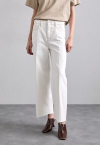 Jeans blancos de pierna ancha con dobladillos desflecados, combinados con una blusa beige y botas de tacón marrones. El material parece de mezclilla con una textura suave.