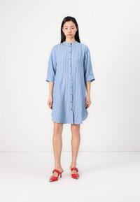 Vestido camiseiro riscado em azul, com colarinho, fecho de botões à frente e bolsos laterais. Combinado com sapatos de salto com tira vermelhos, apresentando acabamentos metálicos.