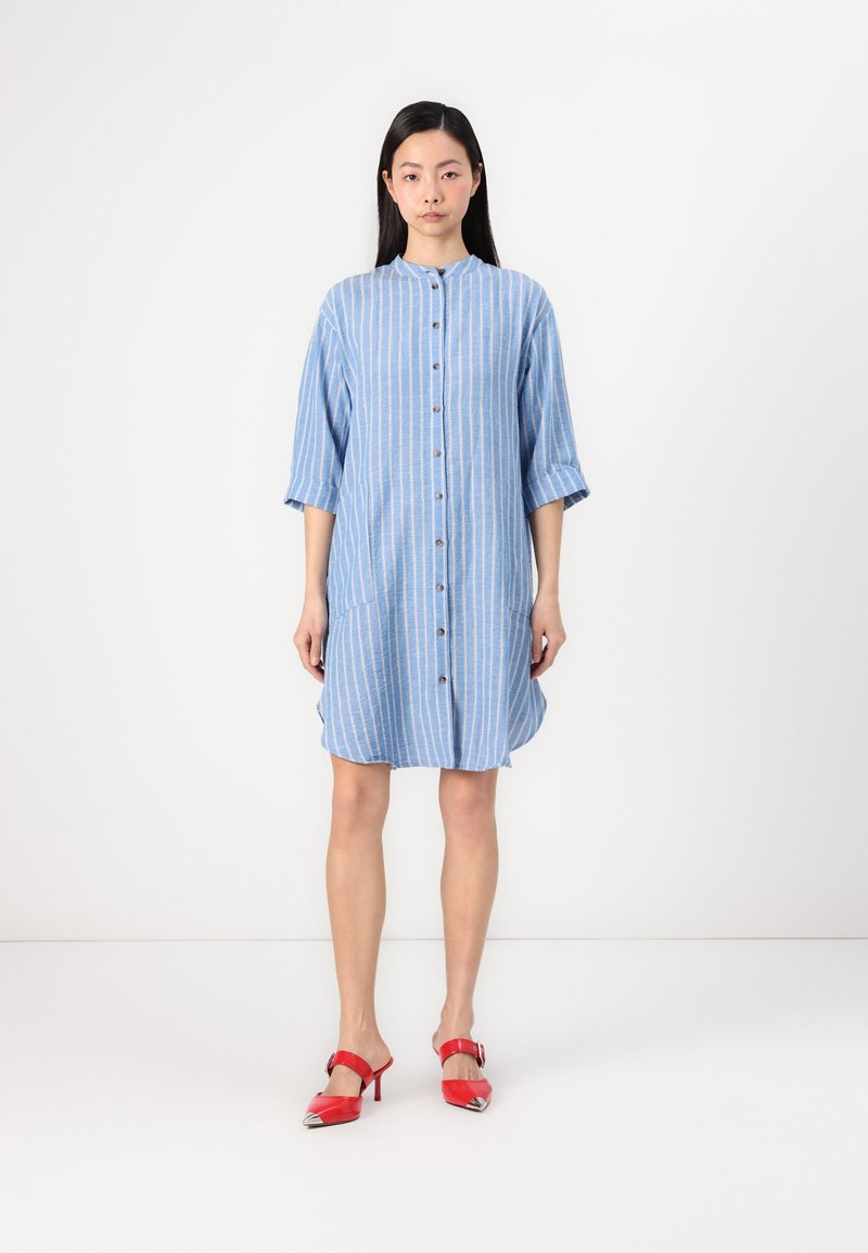 Vestido camiseiro riscado em azul, com colarinho, fecho de botões à frente e bolsos laterais. Combinado com sapatos de salto com tira vermelhos, apresentando acabamentos metálicos.