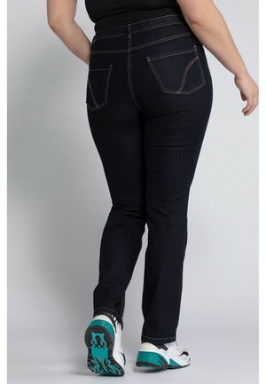 Person trägt dunkelblaue Jeans und weiße Turnschuhe mit türkisfarbenen Sohlen, von hinten vor einem schlichten hellen Hintergrund gezeigt.