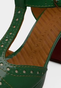 Chaussure en cuir verni vert avec des découpes et des perforations, dotée d'une semelle intérieure marron et d'un talon rouge pour un contraste accru.