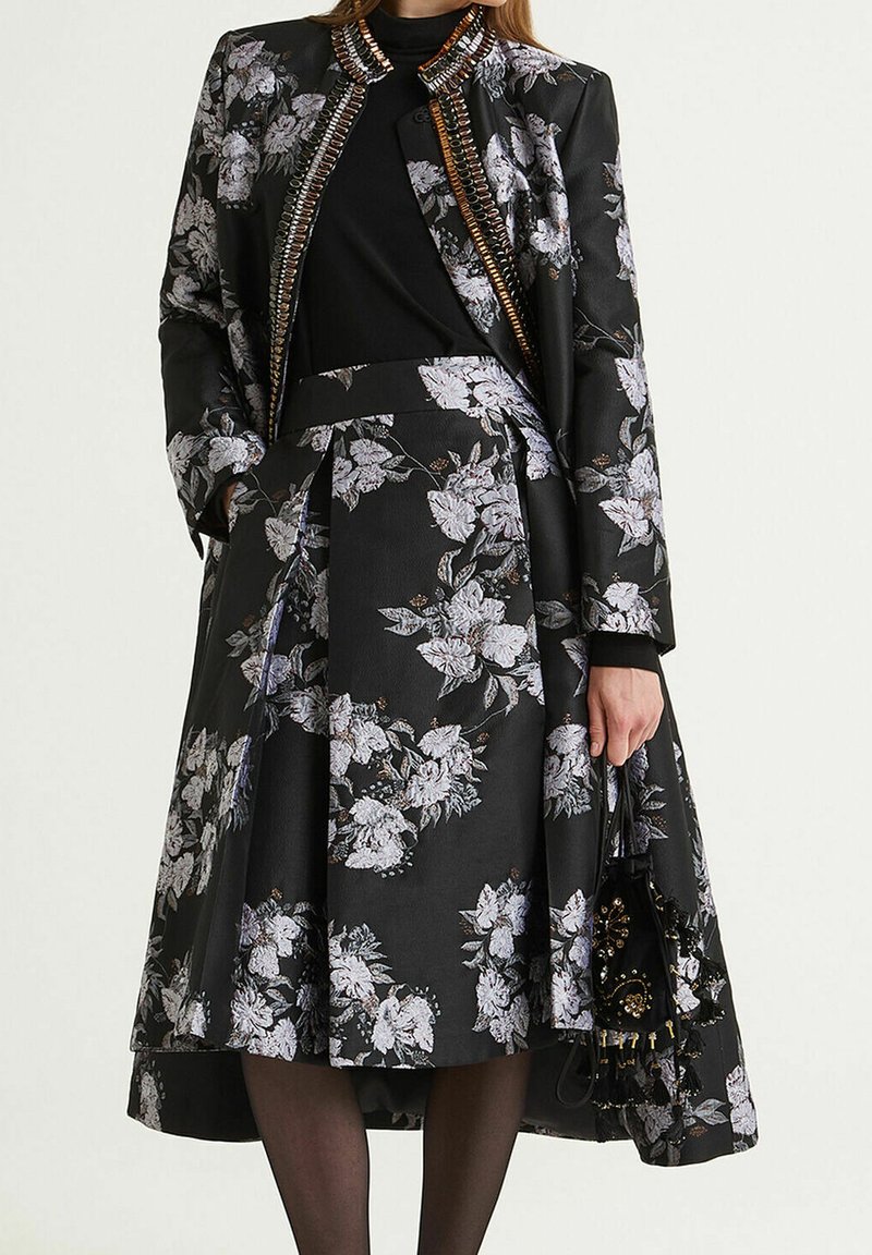 Veste florale noire avec accents en chaîne argentée, assortie à une jupe florale noire. Tissu lisse avec plis structurés. Sac à main noir.