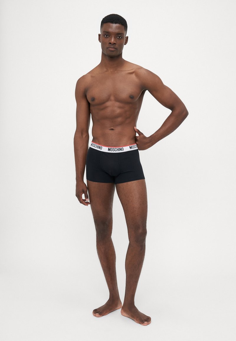Black boxer briefs with a white and red "MOSCHINO" waistband, fitted design, smooth texture, displayed on a model standing barefoot.
