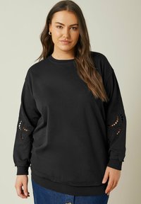 Svart oversized sweatshirt med långa ärmar, med intrikata utskärningsbroderidetaljer vid ärmsluten och en klassisk rund halsringning.