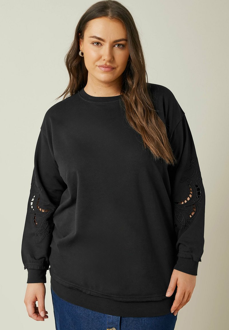 Svart oversized sweatshirt med långa ärmar, med intrikata utskärningsbroderidetaljer vid ärmsluten och en klassisk rund halsringning.