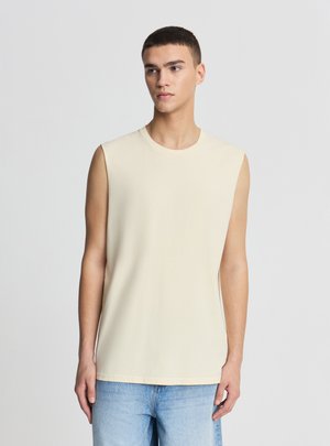 Uomo con maglietta beige senza maniche e jeans azzurri, in piedi davanti a uno sfondo grigio chiaro uniforme, guarda verso destra.