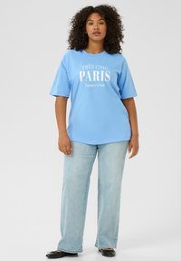 Camiseta de algodón de color azul claro con texto blanco "TRÈS CHIC PARIS Toujours à la mode", combinada con jeans rectos de mezclilla clara y unas bailarinas negras.