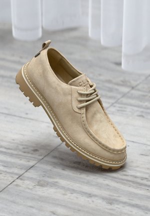 Chaussure décontractée en daim beige avec semelle en caoutchouc épaisse, bout rond et fermeture à lacets, présentée sur un sol en marbre avec des rideaux blancs en arrière-plan.