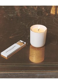 Candela bianca con fiamma in un portacandela di vetro liscio, accanto a una scatola di fiammiferi di legno con un design bianco e nero, su una superficie scura e textured.