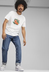 Camiseta blanca con un estampado gráfico colorido en el frente, combinada con jeans de mezclilla azules y zapatillas blancas. Atuendo moderno y casual.