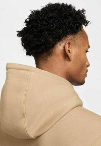 Felpa beige in cotone con un grande cappuccio, dettagli delle cuciture visibili e una texture morbida, vista da dietro, evidenziando i capelli ricci.