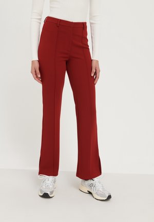 Pantalons rouges évasés en tissu lisse, présentant des détails de couture à l'avant et des fentes sur les côtés à l'ourlet, portés avec des baskets légères.