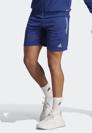 Person trägt blaue Adidas-Shorts, weiße Adidas-Socken und weiße Sneakers und steht auf einem hellen Hintergrund.