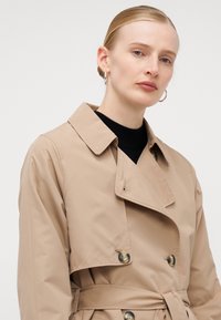 Cappotto beige con design doppiopetto, grandi bottoni e vita regolabile con cintura. Presenta un colletto a punta e una texture di tessuto liscia.