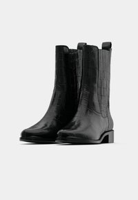 Crne čizme od lažne krokodilske kože do gležnja s okruglim vrhom, kratkim blok petama i trakama za povlačenje na šupljim dijelovima radi lakšeg obuvanja.