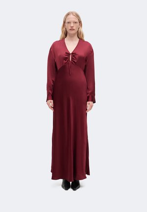 Femme portant une robe en satin bordeaux à manches longues et longueur au sol, avec un détail froncé et un lien au niveau de la poitrine, debout devant un fond uni.