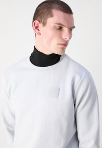 Biela dlhá mikina s dlhým rukávom, s rebrou textúrou, pod ktorou sa nachádza čierny rolák a na hrudi je umiestnená obdĺžniková logo nášivka.