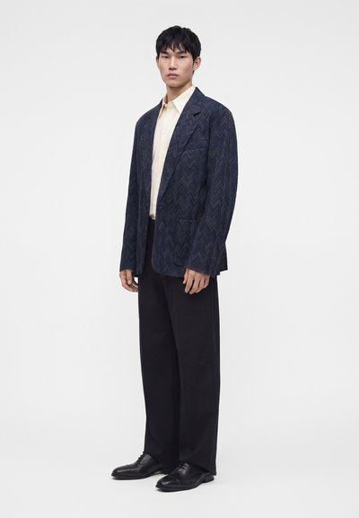 Jeune homme portant un blazer bleu foncé à motifs, une chemise crème, un pantalon noir à jambes larges et des chaussures en cuir noires, debout sur un fond blanc uni.