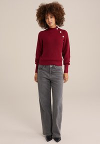 Raudonas raukšlėtas megztinis su aukštu kaklu ir trimis baltais mygtukais ant peties, derinamas su pilkais aukštu liemeniu plačiapadžiais džinsais ir juodais smailiais bateliais.