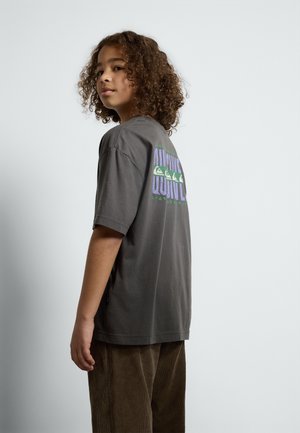 Graues Baumwoll-T-Shirt mit kurzen Ärmeln, das einen bunten Grafikdruck auf dem Rücken zeigt. Kombiniert mit braunen Cordhosen.