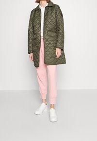 Vadderad olivgrön jacka med krage, med två framtidsfickor och en brun logotypn badge; bärs över rosa sweatpants och vita sneakers.