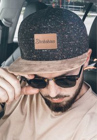 Fekete és szürke foltos sapka barna bőr felirattal, amin a "Blackskies" felirat olvasható, és puha világosszürke suede karimával. Kerek fekete napszemüveg és bézs póló.