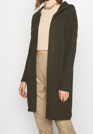 Kobieta ubrana w teksturowany ciemnozielony kardigan z kapturem, nałożony na beżowy, skrócony sweter prążkowany, oraz jasnobeżowe spodnie, ozdobiona złotym naszyjnikiem z łańcuszkiem.