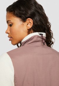 Gilet sans col présentant une coque mauve clair et une doublure en polaire blanche. Comprend un ajustement à bascule au niveau du cou pour un maintien sécurisé.