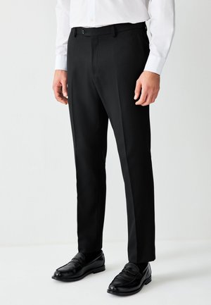 Mand iført sorte dressbukser, sorte sko og en hvid langærmet skjorte med knapper, der står mod en ensfarvet baggrund.