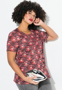 Camisa de manga corta en patrones rojo y negro con diseño de labios; modelo sosteniendo un bolso de mano metálico plateado en forma de labios; jeans grises.