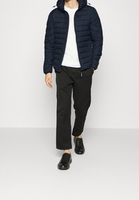 Mann, der mit einer marineblauen gesteppte Jacke, einem weißen T-Shirt, schwarzen Hosen und schwarzen Sneakers vor einem hellen Hintergrund geht.