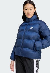 Marineblauwe pufferjack met hoge kraag, gequilte ontwerp en wit Adidas-logo op de borst. Beschikt over een voorsluiting met rits en elastische manchetten.