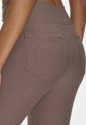 Nahaufnahme einer Person, die hochtaillierte braune Leggings mit aufgesetzten Gesäßtaschen und sichtbaren Nahtdetails trägt.