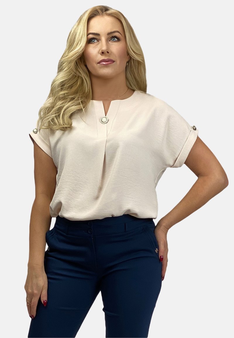 Bluzka w jasnobeżowym materiale o luźnym kroju, z krótkimi podwiniętymi rękawami, plisą z przodu oraz dekoracyjnymi guzikami. Zestawiona z granatowymi spodniami.