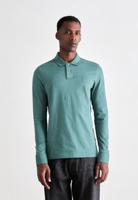 Cămașă polo cu mânecă lungă, culoare teal, material din amestec de bumbac, guler cu nasturi, detaliu mic cu logo pe piept, asortată cu pantaloni din denim închisi la culoare.