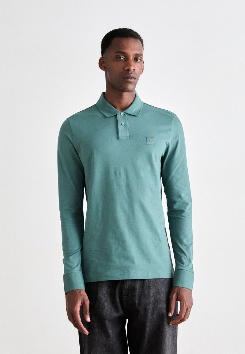 Cămașă polo cu mânecă lungă, culoare teal, material din amestec de bumbac, guler cu nasturi, detaliu mic cu logo pe piept, asortată cu pantaloni din denim închisi la culoare.