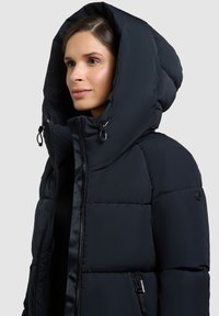 Giacca a vento in navy, con un design spesso e trapuntato. Presenta un grande cappuccio staccabile e tasche con zip con accenti in metallo nero.