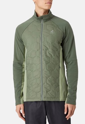 Man wearing olive green quilted jacket with hexagon pattern, zip front, and long sleeves over black pants on white background.