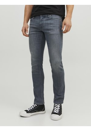Mand iført slim-fit falmet grå jeans og sorte høje sneakers med hvide snørebånd og såler, står mod en ensfarvet baggrund.